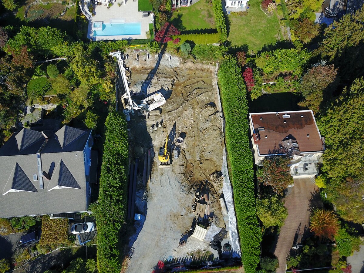 Aerial view of residential construction site showing pile driving equipment and deep excavation with comprehensive shoring system in West Vancouver neighbourhood