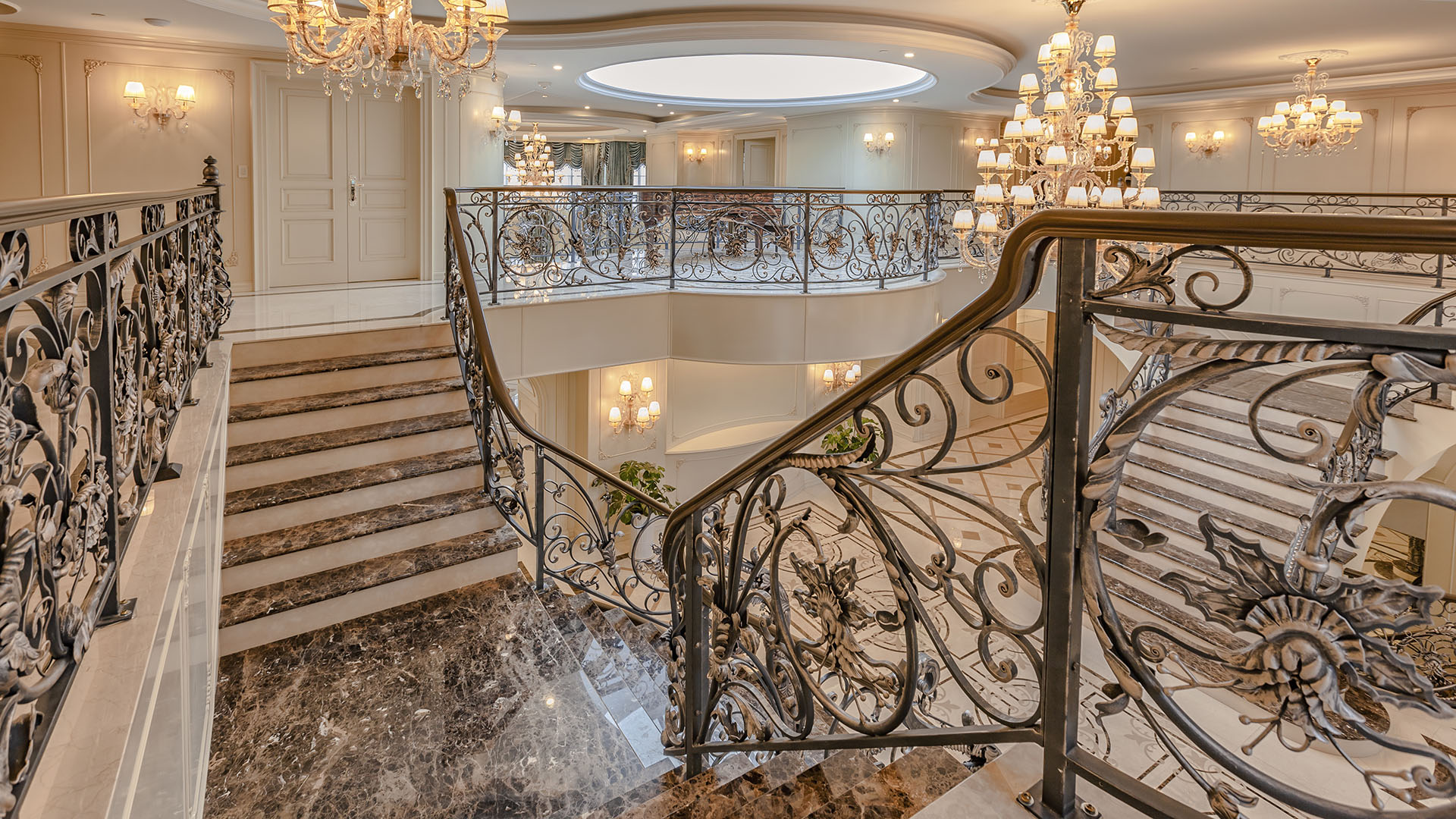 Ambleside Palace upper landing view of dual marble staircases with ornate iron railings and crystal chandeliers in West Vancouver