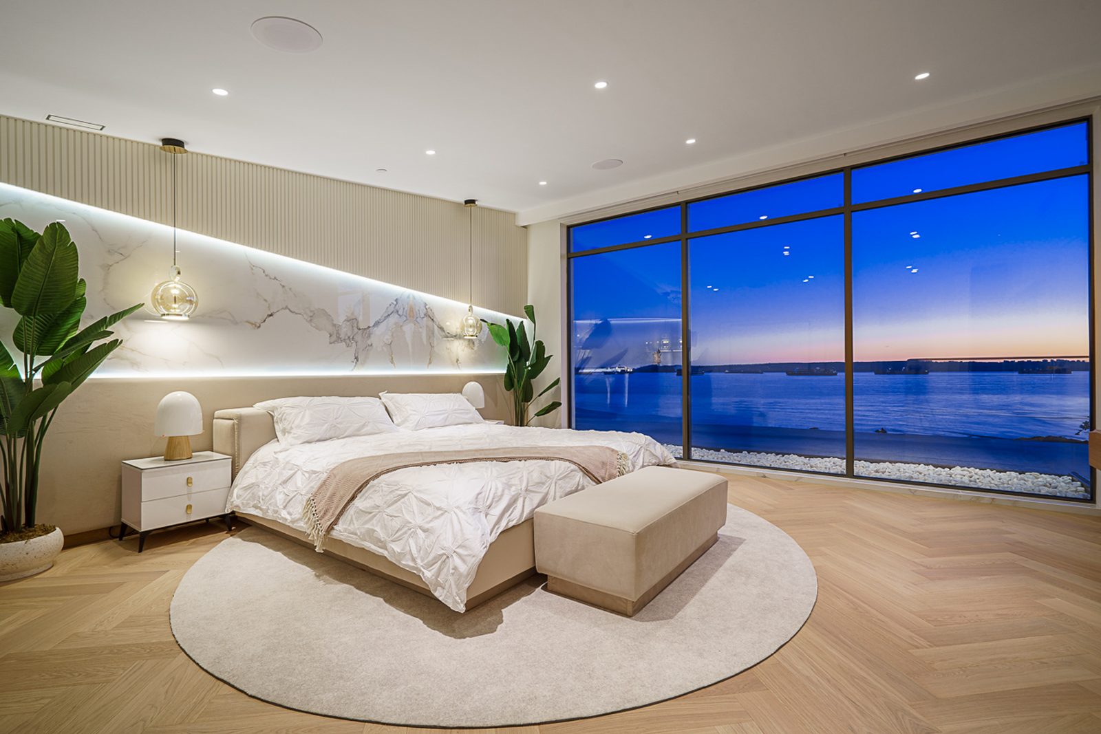 Master bedroom at dusk with harbour lights through floor-to-ceiling glass and cove lighting West Vancouver