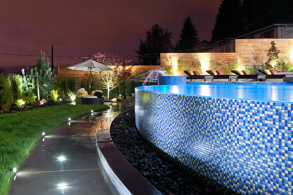 Infinity edge glass-tiled swimming pool at twilight with illuminated waterfall jets loungers and tiered landscaping Millstream British Properties West Vancouver luxury home by Eurohouse