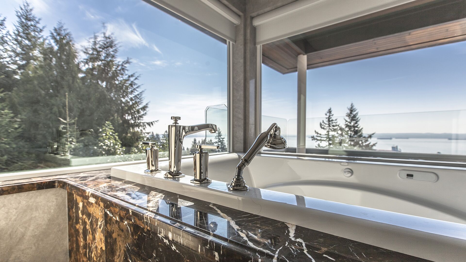 Close-up of polished nickel tub fixtures on Emperador Dark marble surround with ocean treetop and sky views through corner window at Rockridge Classic built by Eurohouse Construction