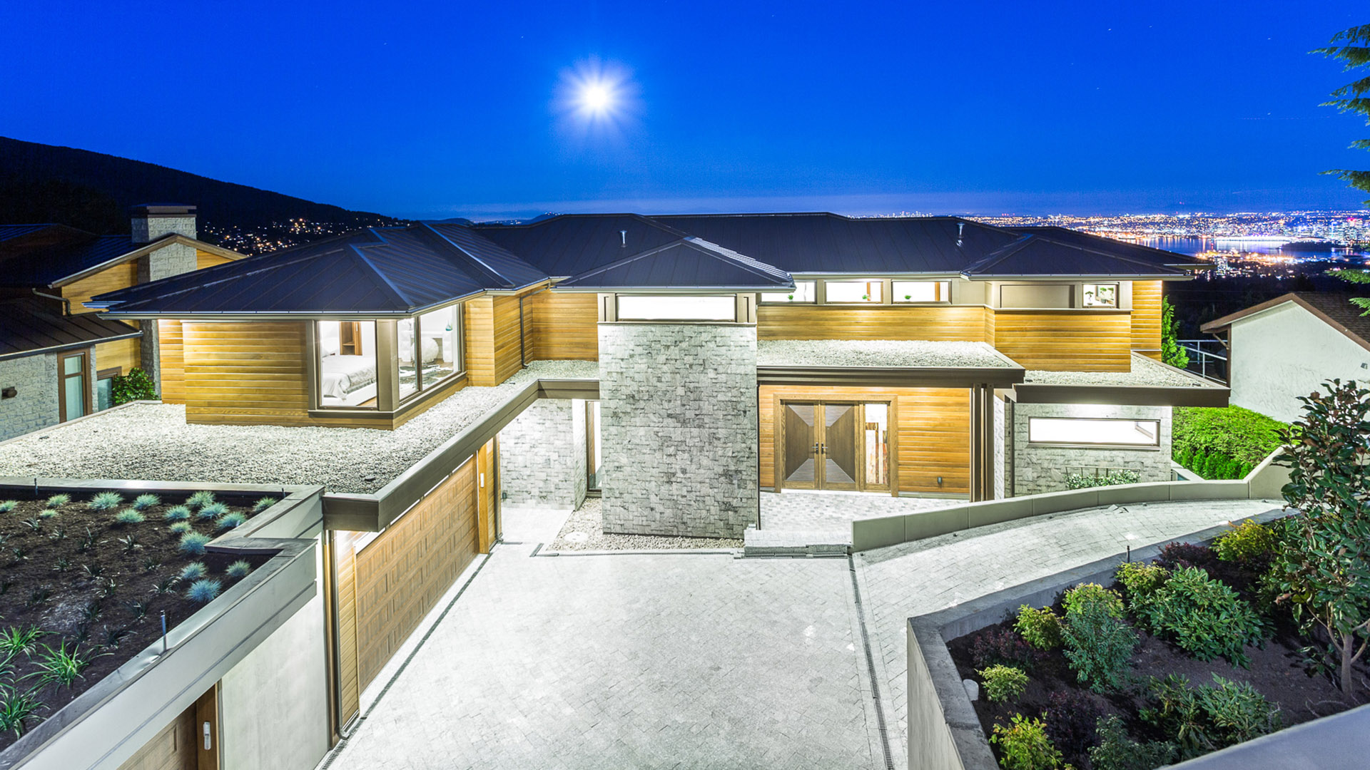 Front entrance of West Coast Contemporary at twilight showing granite facade cedar garage doors metal roof and LED-lit driveway with city lights in background British Properties West Vancouver