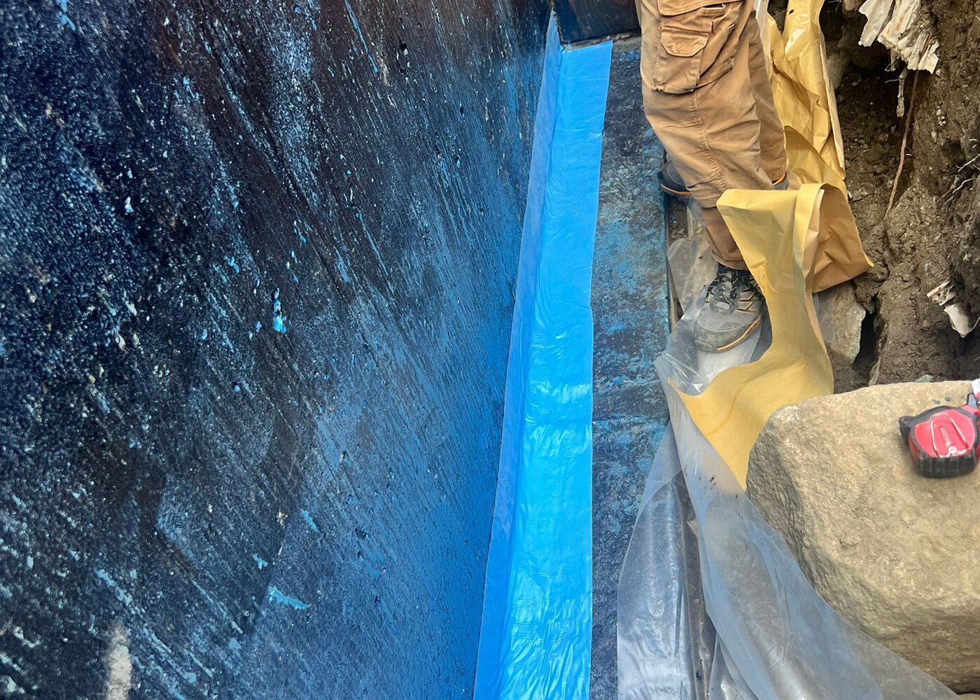 Worker applying Blueskin waterproofing membrane to concrete foundation wall in deep excavation trench