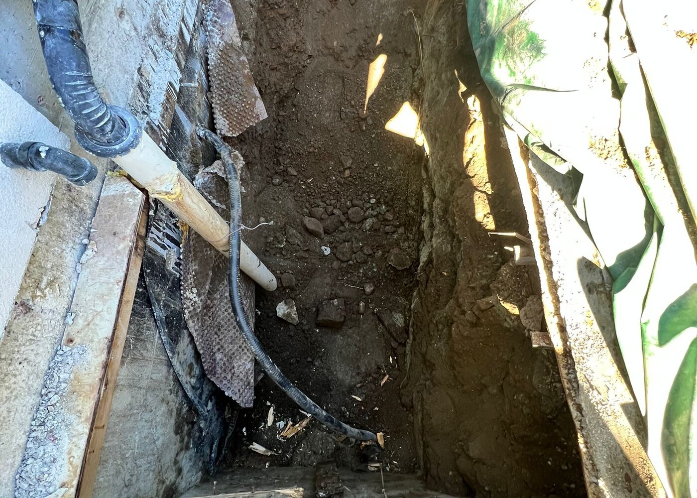 Deep excavation pit exposing foundation footing, utilities, and failed waterproofing membrane requiring replacement