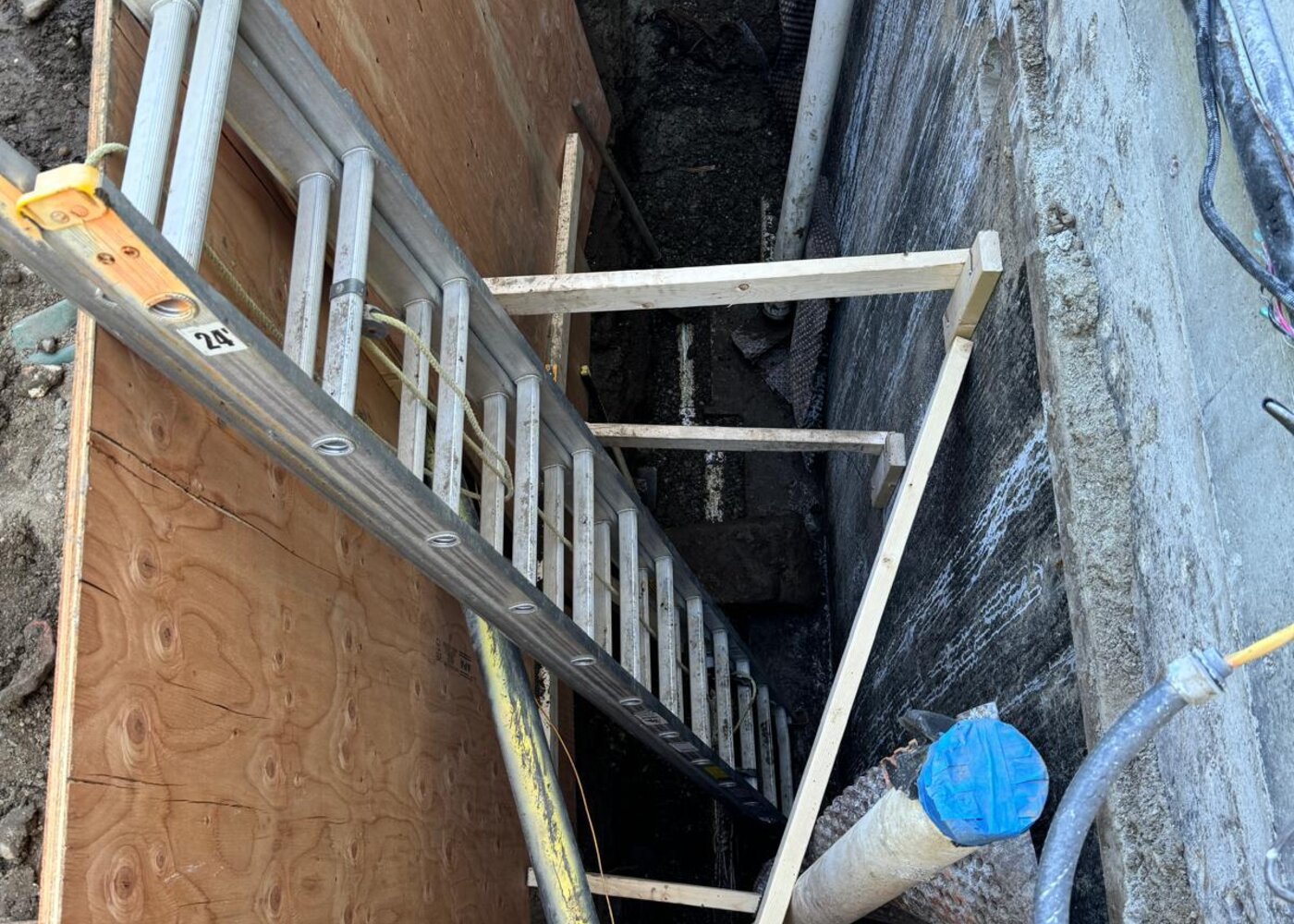 Ladder access into deep foundation repair trench with exposed utilities and shoring during home warranty excavation