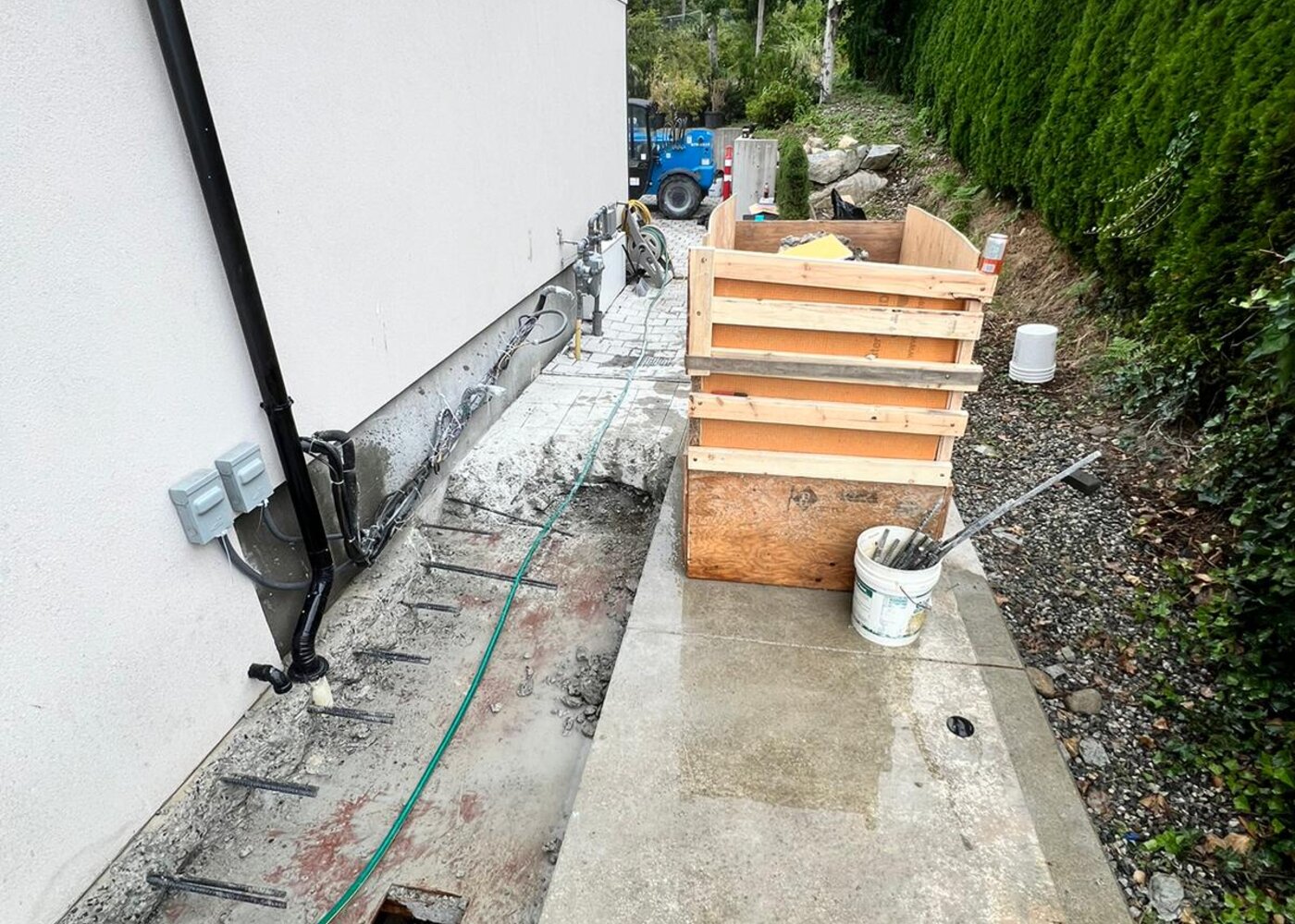 Excavated foundation trench along exterior wall showing exposed rebar, formwork, and drainage opening during home warranty repair in West Vancouver
